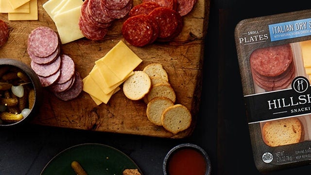 Overhead view of Italian dry salami, sliced cheeses, crackers and olives on a board beside Hillshire Small Plates Italian Salami packaging