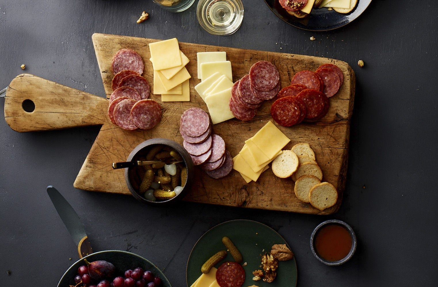 Overhead view of a rustic wooden charcuterie board with sliced salami, assorted cheeses, pickles, and toasted bread on a dark table
